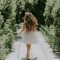 Back view of a woman in a sunlit park walking amidst lush green trees on a paved pathway.